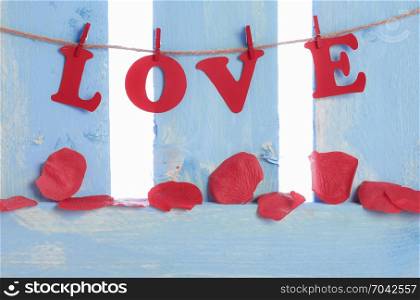 Red rose petals made of soap, spread on a blue wooden fence and the word love spelled with red paper letters and tied to a string with wooden clips.