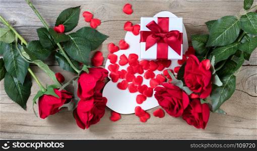 red roses surrounding white gift box and many hearts on weathered wooden background in flat lay view