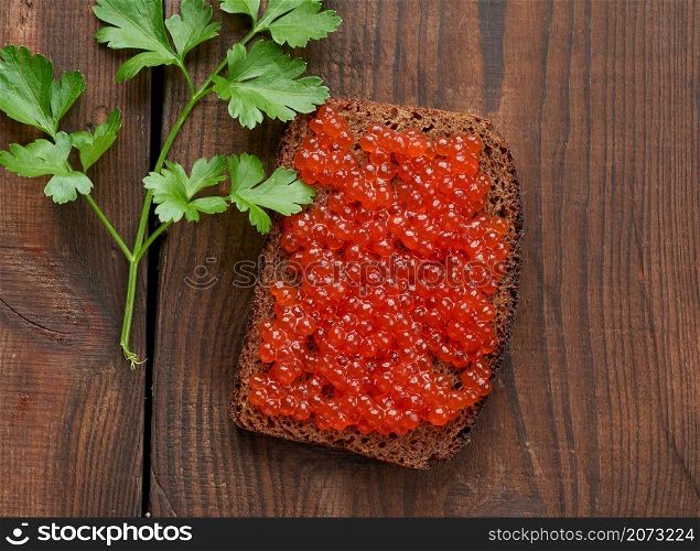 red salmon caviar lies on a piece of rye bread. Brown wooden table, top view