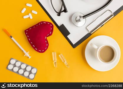 red stitched heart coffee cup stethoscope eyeglass injection blister packed medicine clipboard against yellow backdrop