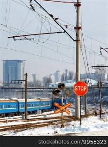 Red stop sign at a railway crossing against the background of railway tracks, blue train carriages and cityscape at the entrance to the city, winter cityscape, vertical image.. Red stop sign at the railway crossing at the entrance to the city against the background of the winter season.