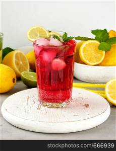 red strawberry lemonade in a glass on a round white wooden board, yellow lemons are behind