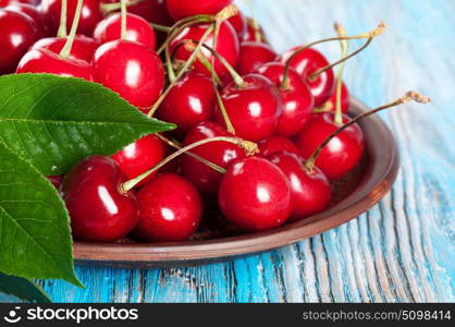 Red sweet cherry and green leaves in a clay dish on a blue wooden background