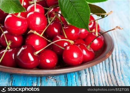 Red sweet cherry and green leaves in a clay dish on a blue wooden background