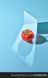 Red tomato with a green stem and a mirror on a blue background with shadows and a copy of a spacer. Creative food composition. Composition from a mirror and a ripe tomato on a blue background with reflection of shadows and copy space. Organic vegetable