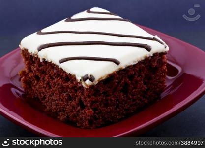 Red velvet cake with white icing and drizzles of chocolate placed on square, red plate on black background.