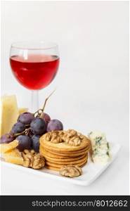 Red wine, different varieties of cheese, grapes, crackers and walnuts on the white porcelain plate on white background