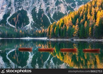 Red wooden boats on Braies lake at sunrise in autumn in Dolomites, Italy. Landscape with fall forest, mountains, water with reflection, trees with colorful foliage. Travel in italian alps. Nature