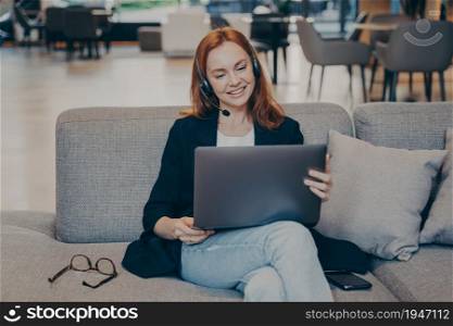 Redhead attractive smiling female in earphones headset and laptop watching online webinar in internet or enjoying e-learning, sitting on cozy sofa in coffee shop. Distance education concept. Redhead smiling female blogger sitting on cozy sofa in cafe and watching online webinar on laptop