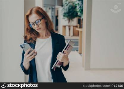 Redhead business woman with serious look in eyewear using cell phone, checking email or newsfeed of social network account, holding laptop and notebook, looking at smartphone while standing in office. Redhead business woman with serious look in eyewear using cell phone while standing in office