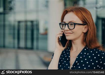 Redhead businesswoman has telephone conversation uses modern cellular gadget for consultancy discusses organisation planning phones via mobile application wears spectacles polka dot clothes.. Redhead businesswoman has telephone conversation uses modern cellular gadget