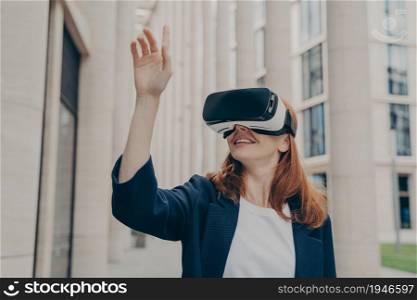 Redhead businesswoman managing business project through virtual reality platform. Smiling young female office worker enjoying spending time with VR headset glasses, pointing with forefinger up in air. Redhead businesswoman managing business project through virtual reality platform
