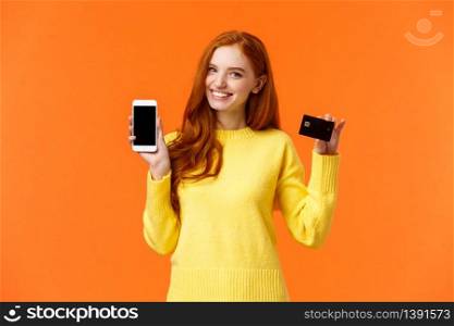 Redhead caucasian girl showing application on smartphone as holding mobile phone and credit card, using mobile banking app, cheerfully smiling camera, advertising over orange background.. Redhead caucasian girl showing application on smartphone as holding mobile phone and credit card, using mobile banking app, cheerfully smiling camera, advertising over orange background