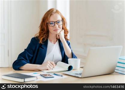 Redhead curly woman office worker analyzes data, makes accounting report, poses in coworking office, improves online sales, drinks coffee, wears spectacles and formal wear. Female employee indoor
