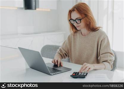 Redhead European female accountant analyzes documents uses calculator makes report uses laptop computer sits at white desktop wears spectacles beige jumper poses against white home interior.. Redhead European female accountant analyzes documents uses calculator makes report