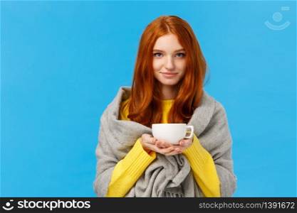 Redhead girl trying get warm, feeling cold wrapping herself with scarf on shoulders, drinking hot tea, holding teapot and smiling lovely, standing tender and cute blue background. Copy space. Redhead girl trying get warm, feeling cold wrapping herself with scarf on shoulders, drinking hot tea, holding teapot and smiling lovely, standing tender and cute blue background
