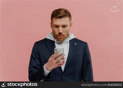 Redhead guy in jacket, hoodie, reading unbelievable news on phone, stunned, standing alone against pink wall