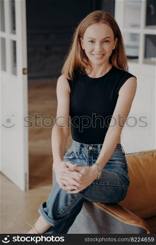 Redhead lady in jeans and bodysuit is smiling. Cheerful stylish lady is sitting on an armchair armrest. Portrait of glamorous slim fair young woman. Happy european woman in apartment.. Redhead lady in jeans and bodysuit is smiling. Cheerful stylish lady is sitting on armchair armrest.