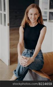 Redhead lady in jeans and bodysuit is smiling. Cheerful stylish lady is sitting on an armchair armrest. Portrait of glamorous slim fair young woman. Happy european woman in apartment.. Redhead lady in jeans and bodysuit is smiling. Cheerful stylish lady is sitting on armchair armrest.