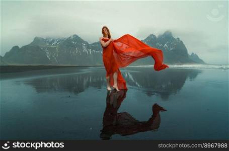 Redhead model in red dress on Reynisfjara beach scenic photography. Picture of person with hills on background. High quality wallpaper. Photo concept for ads, travel blog, magazine, article. Redhead model in red dress on Reynisfjara beach scenic photography