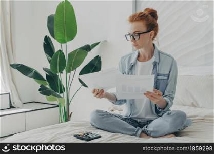 Redhead serious Caucasian woman sits in lotus pose on bed studies documents calculates bills makes financial report at home enjoys cozy domestic atmosphere takes care of budget count expenses. Redhead serious Caucasian woman sits in lotus pose on bed studies documents calculates bills