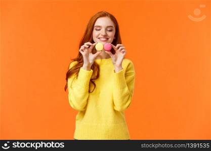 Redhead silly and happy gorgeous girl buy tasty macarons, holding food and looking with desire and affection, like sweets, want eat them, standing orange background satisfied and joyful.. Redhead silly and happy gorgeous girl buy tasty macarons, holding food and looking with desire and affection, like sweets, want eat them, standing orange background satisfied and joyful