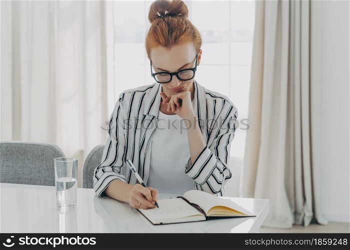 Redhead woman in striped shirt takes notes writes down information in notebook counts month spendings focused in notebook sits at table drinks fresh water cozy domestic interior plans schedule. Redhead woman in striped shirt takes notes writes down information in notebook