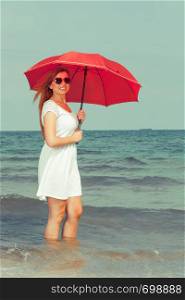 Redhead woman wearing gauzy white dress walking on beach holding red umbrella, having nice relaxing summertime walk. Redhead woman walking on beach holding umbrella
