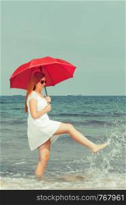 Redhead woman wearing gauzy white dress walking on beach holding red umbrella, having fun playing with water. Redhead woman walking on beach holding umbrella