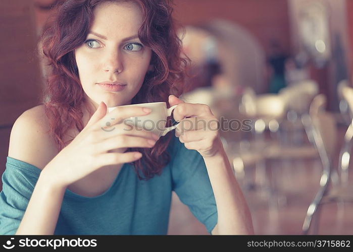 Redhead women sitting in the cafee and holding cup of coffee in the hands looking away