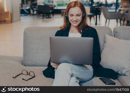 Redhead young woman communicating with clients using laptop and wireless headphones, smiling lady talking during virtual call sitting on sofa in cafe shop. Online education and remote working concept. Redhead young woman communicating with clients using laptop and wireless headphones in cafe shop