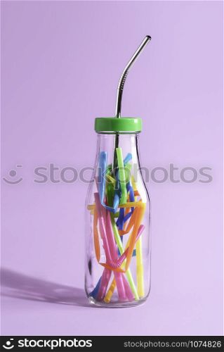 Reduce plastic pollution concept with a reusable metal straw in a glass bottle full of colorful plastic, on purple paper. Eco-friendly drinking straw.