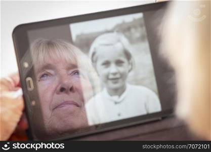 Reflection of a senior caucasian woman looking at old photos of herself on a tablet computer themes of contrasts the ageing process nostalgia and childhood