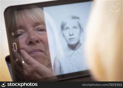 reflection of a senior caucasian woman looking at old photos of herself on a tablet computer themes of contrasts the ageing process nostalgia and childhood