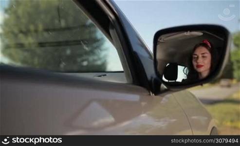Reflection of beautiful brunette woman in side rearview mirror of car. Sensual young female looking at the car rearview mirror. Pretty girl staring at car&acute;s side rearview mirror with toothy radiant smile. Slow motion. Steadicam stabilized shot.