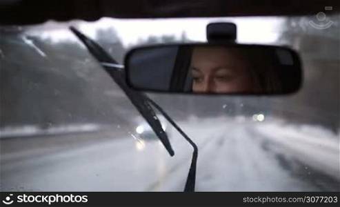 Reflection of beautiful woman driving car in wintertime in the rearview mirror. The face of young blond female in the rear view mirror while driving automobile during snow on highway. View from inside of car.