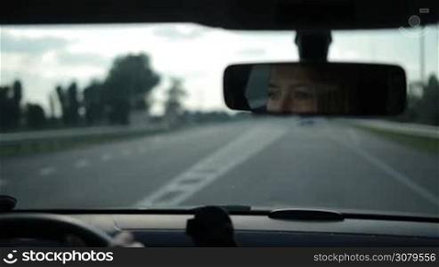 Reflection of charming blond female driver with amazing blue eyes in car rear-view mirror while driving vehicle on highway. Young woman driving car on freeway and looking at rear-view mirror.