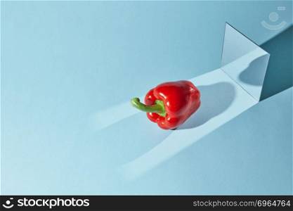 Reflection of shadow from pepper in a mirror. Diagonal long shadows from pepper and mirror on the table surface.. Red pepper, a square mirror on a blue background, a reflection of an pepper and from shadow in a mirror.
