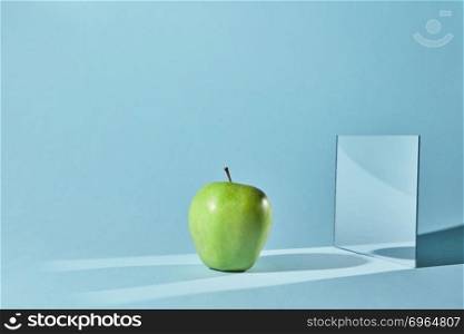 Reflection of the shadow from apple in a mirror. Diagonal long shadows from apple and mirror on the blue table surface.. The apple is green, a square mirror on a blue background, a reflection of an apple and from shadows in a mirror.
