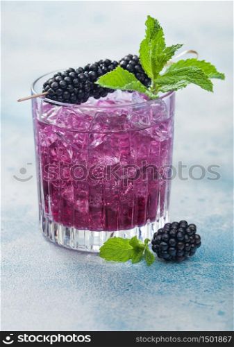 Refreshing summer cocktail with blackberry in crystal glass with ice cubes and mint on blue background. Soda and alcohol mix. Macro