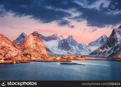 Reine village and snow covered mountains at beautiful sunrise in winter. Lofoten islands, Norway. Landscape with blue sea, snowy rocks, houses and traditional rorbus, colorful sky with clouds. Coast
