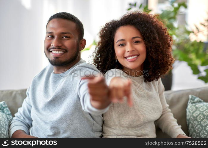relations and people concept - happy african american couple sitting on sofa and pointing to camera at home. happy african american couple pointing to camera