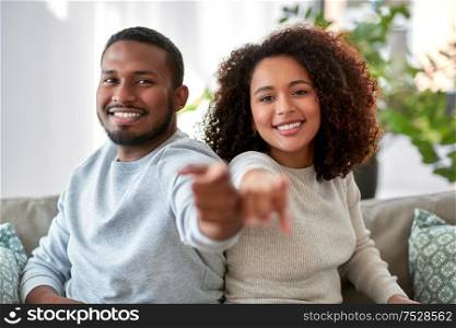 relations and people concept - happy african american couple sitting on sofa and pointing to camera at home. happy african american couple pointing to camera