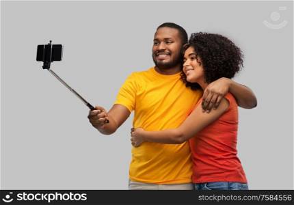 relationships and people concept - happy african american couple taking picture by smartphone on selfie stick over grey background. african american couple taking selfie by cellphone
