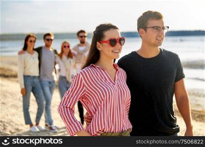 relationships and people concept - happy couple with group of friends hugging on beach in summer. happy friends walking along summer beach