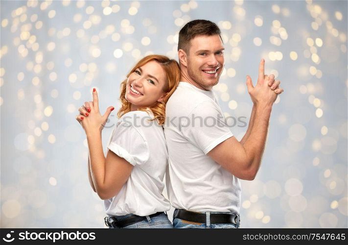 relationships and people concept - portrait of happy couple in white t-shirts making gun gesture over holidays lights background. couple in white t-shirts shirts making gun gesture