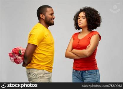 relationships, couple and people concept - happy smiling african american man hiding bunch of flowers behind his back from displeased woman over grey background. man hiding bunch of flowers behind back from woman