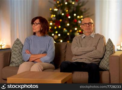 relationships difficulties, problem and people concept - unhappy senior couple sitting on sofa at home over christmas tree lights on background. unhappy senior couple at home on christmas