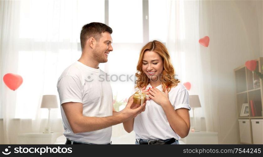 relationships, holiday and valentine&rsquo;s day concept - happy couple in white t-shirts with gift over bedroom decorated with heart shaped balloons background. happy couple with gift on valentines day at home