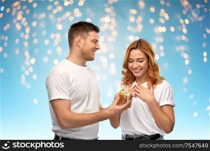 relationships, holiday and valentines day concept - happy couple in white t-shirts with christmas gift over lights on blue background. happy couple in white t-shirts with christmas gift